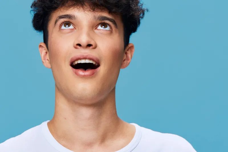 young man with curly hair looking up with surprised expression on blue background wearing white shirt, capturing emotion and youth vitality
