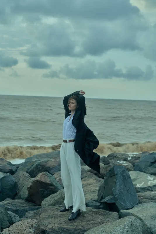 Woman stands on rocky seashore by the sea, wind lifting a dark coat and flowing outfit, arms raised, eyes toward the horizon, dramatic fashion moment against cloudy sky and water