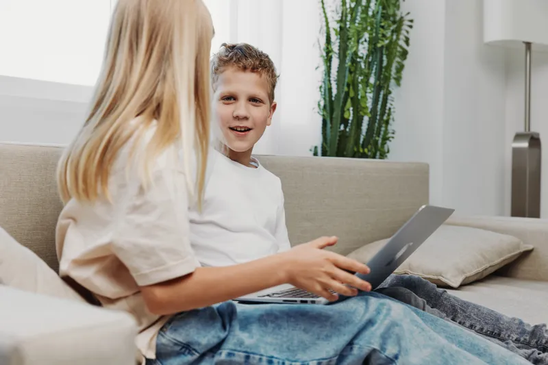 Two children sitting on a sofa using a laptop together, engaged in conversation in a bright living room with natural light and indoor plants. Concept of learning, technology, and childhood
