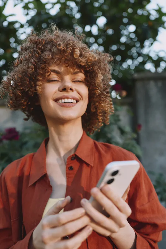 A smiling woman outdoors in casual attire uses a smartphone, enjoying a sunny day. This lifestyle portrait highlights confidence, connection, and everyday moments
