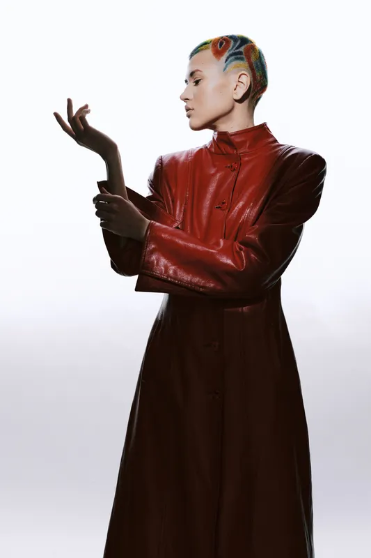 Woman with colorful hair and red leather coat poses confidently against a bright white background, showcasing modern fashion and artistic style with a minimalist vibe.
