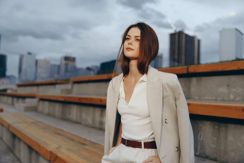 Young woman in business outfit with beige blazer and white shirt standing outdoors on urban staircase during cloudy day