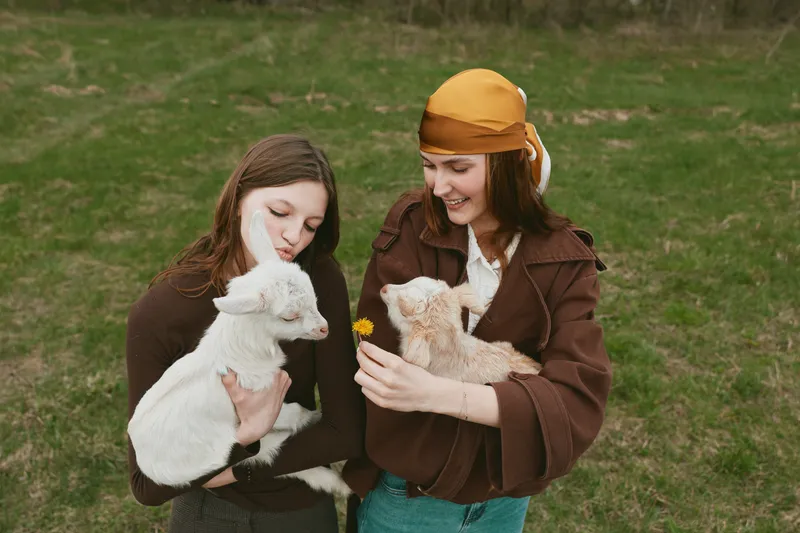Two women hold a young goat in a sunny rural setting, sharing a tender moment. A wholesome lifestyle concept highlighting animal companionship, care, and everyday farm life.