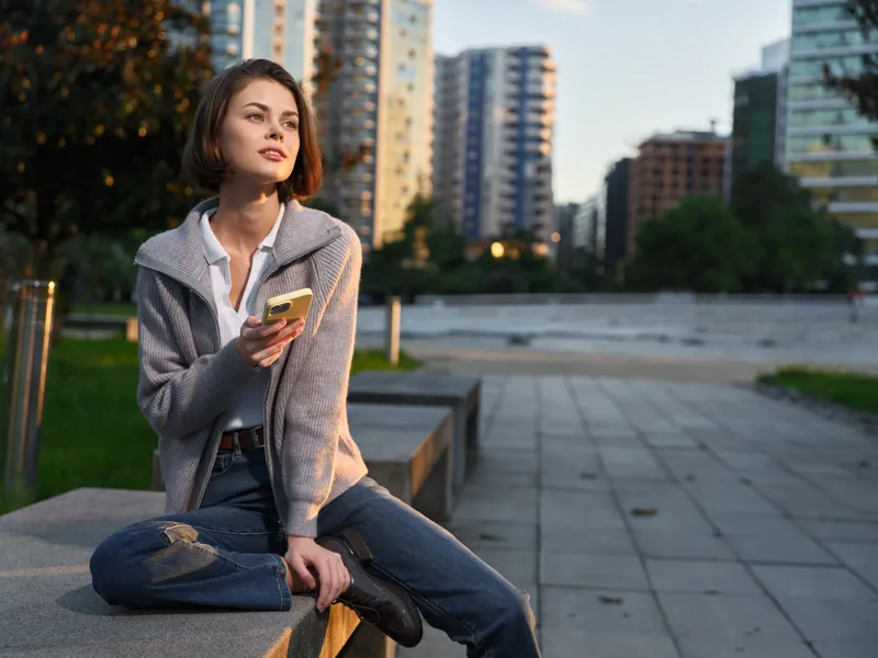 A young woman sits on a concrete ledge in a modern city park, texting on a mobile device. She wears a casual jacket and jeans, appearing focused and relaxed.