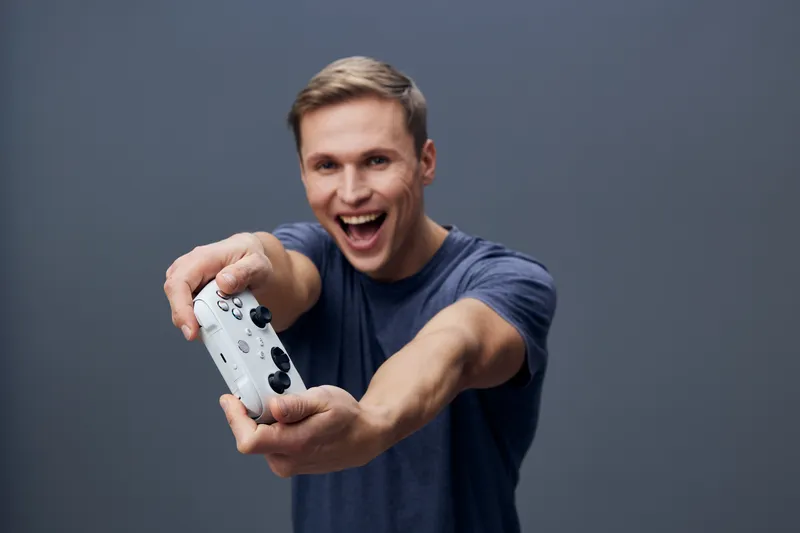 Smiling man with short hair and casual blue t-shirt holding a video game controller, looking excited and engaging with the camera, standing against a plain gray background, studio portrait, people lif