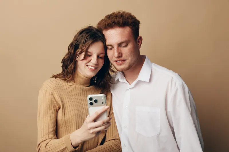 Young couple looking at smartphone together smiling, standing close on beige background. Concept of technology, relationship, communication, and modern lifestyle.