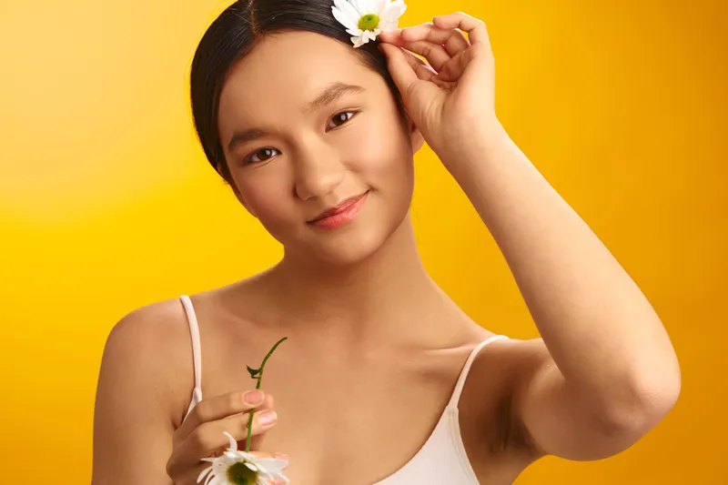 Asian woman holds a flower near her hair, standing against a bright yellow background, smiling softly to convey a fresh, approachable portrait with warm, inviting tones.