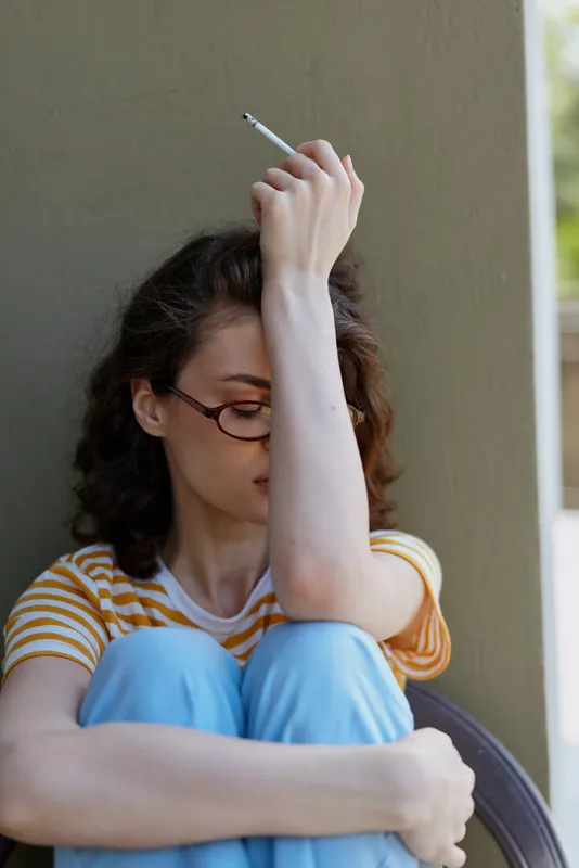 Woman lifestyle home, a reflective mood as a young woman sits against a wall with knees up, holding a cigarette, wearing glasses and a striped shirt with blue pants, conveying solitude.