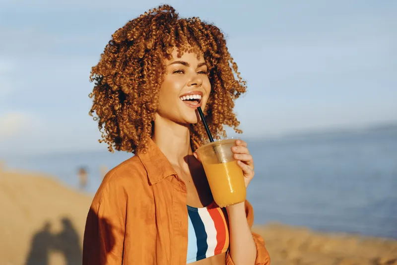 Smiling woman with curly hair holding a glass of iced coffee with straw on a sunny beach, summer vacation mood, warm light, relaxed outdoor lifestyle close to the ocean view
