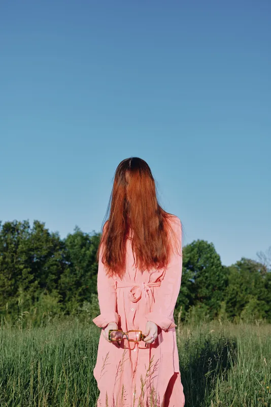 A young woman in a pink dress stands in a lush green field against a clear blue sky, embodying a serene and contemplative mood while holding sunglasses