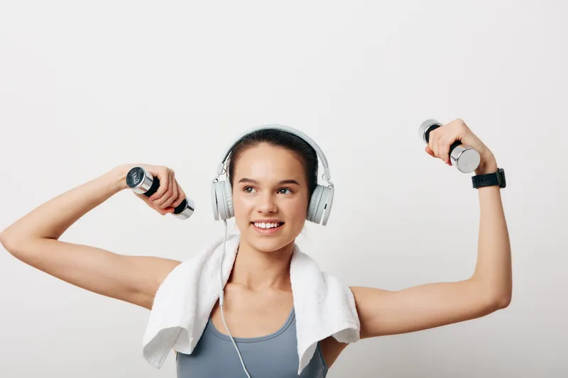 Woman fitness enthusiast lifts dumbbells with arms raised, headphones on and a towel draped, in a bright studio during a workout, showcasing strength, focus, and athletic routine.