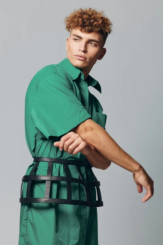 Young man with curly hair wearing green outfit and black cage belt posing with crossed arms on gray background in studio fashion portrait.