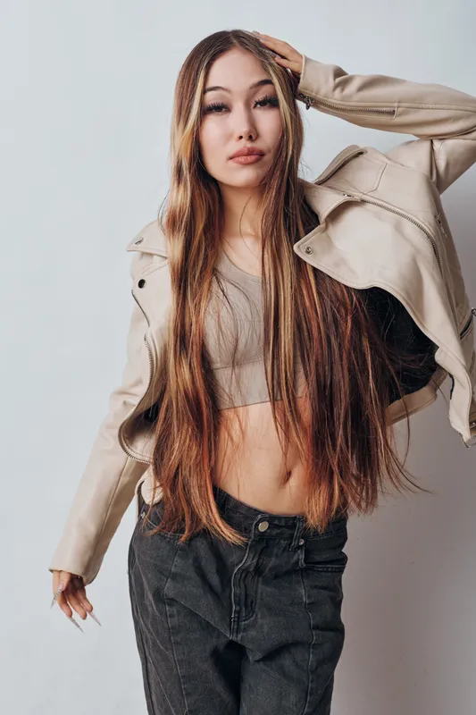 Young woman with long straight hair wearing casual beige jacket and crop top posing against plain background. Fashion model with confident expression and relaxed pose in studio setting.