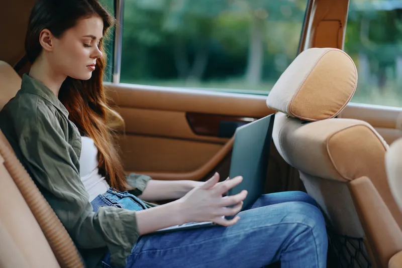 Young woman working on a laptop in the back seat of a car, expressing concentration and focus, dressed casually in a jacket, with a natural green background.