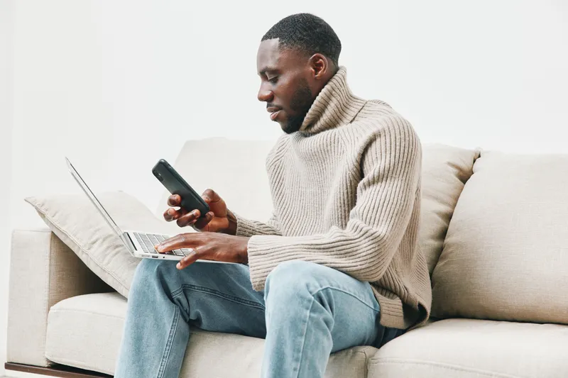 Focused man using laptop and smartphone at home while sitting on couch, wearing cozy sweater and jeans, showcasing a relaxed and modern lifestyle