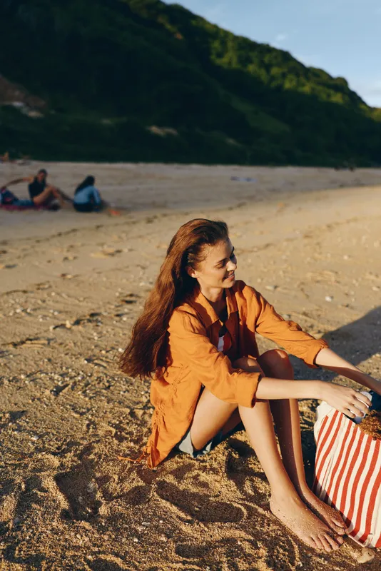 Relaxing woman sitting on sandy beach in warm sunlight, wearing orange dress and striped skirt, holding hair, smiling, peaceful travel mood with green forested mountains in background.