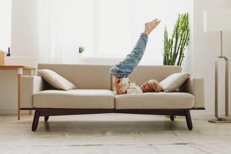 Young child lying upside down on beige sofa in bright living room, wearing casual jeans and white shirt. Concept of playful childhood, home comfort, and relaxation indoors.