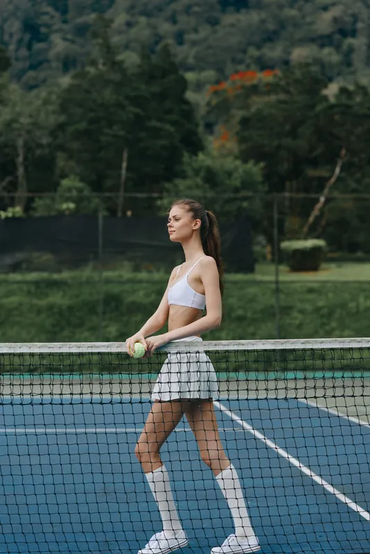 Young female tennis player in a sporty outfit with a confident expression on a tennis court surrounded by greenery, showcasing athleticism and freshness