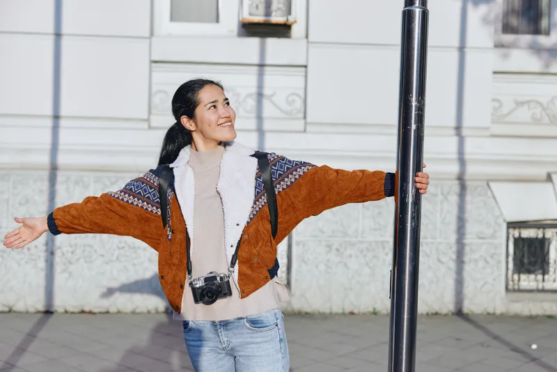 Woman with open arms on a sunny street, wearing a brown jacket and denim, posing beside a pole for a fashion and lifestyle shot, with an urban backdrop and confident expression