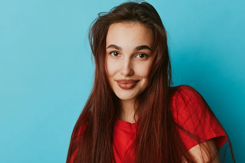 Young woman with long hair and red shirt smiling at camera against blue background. Portrait of happy female with natural makeup and casual style in studio lighting.