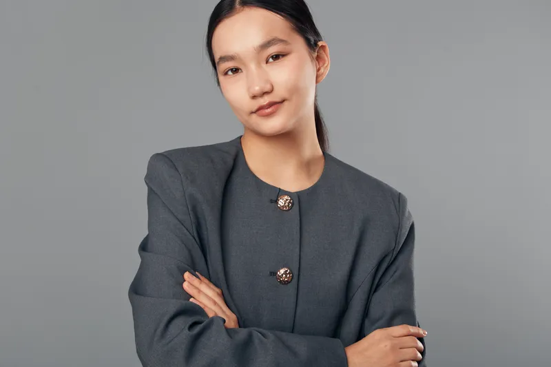 Young confident businesswoman standing with arms crossed in formal suit on gray background. Professional Asian woman posing in studio with calm expression and neat hairstyle.