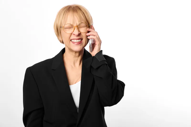 Smiling elderly business woman in black suit, holding phone to her ear, wearing glasses, white background, cheerful call concept, friendly communication, corporate