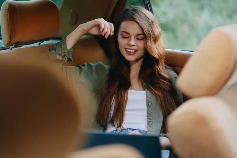 A young woman in casual attire relaxes in a car, enjoying her time while using a laptop, showcasing a tranquil outdoor atmosphere and warm tones.