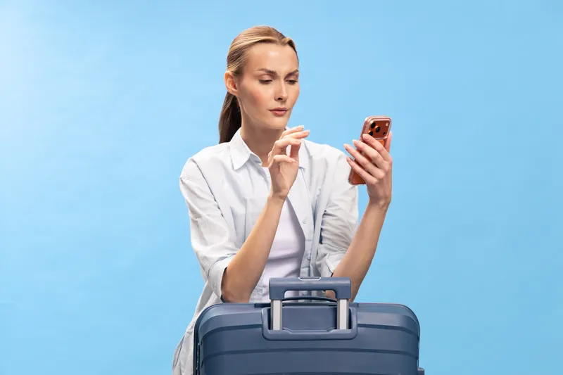 Young woman in white shirt uses smartphone sitting on blue suitcase against blue background. Concept of travel, communication, and modern lifestyle with casual outfit and focused expression.