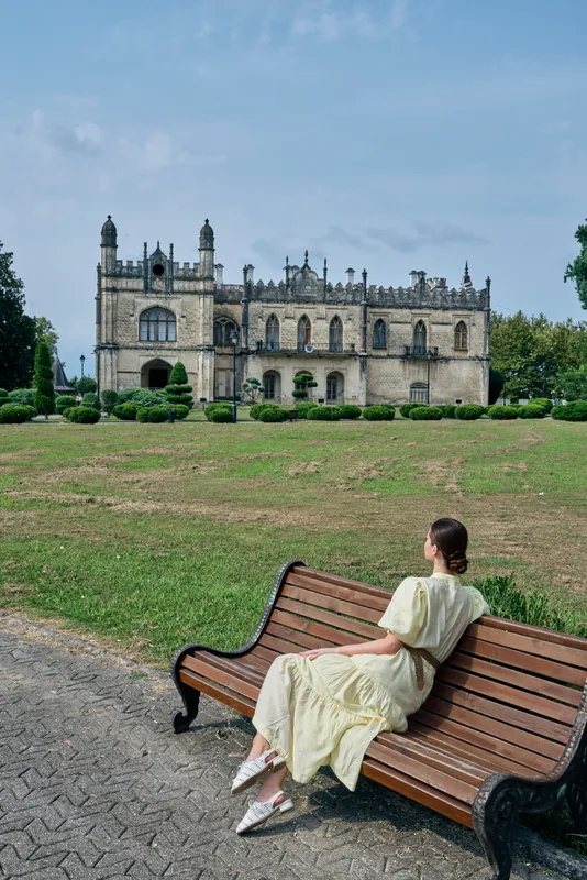 Woman on bench facing historic mansion in park for authenticity inclusivity, vintage dress solitude