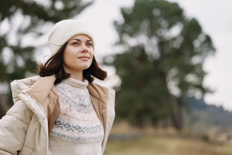 Woman in a warm beanie and light coat stands outdoors in a windswept field, calm and thoughtful as trees blur in the background against a soft cloudy sky.