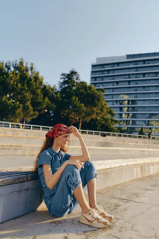 young woman sitting outdoors wearing denim outfit and red bandana looking thoughtful in sunny urban park setting