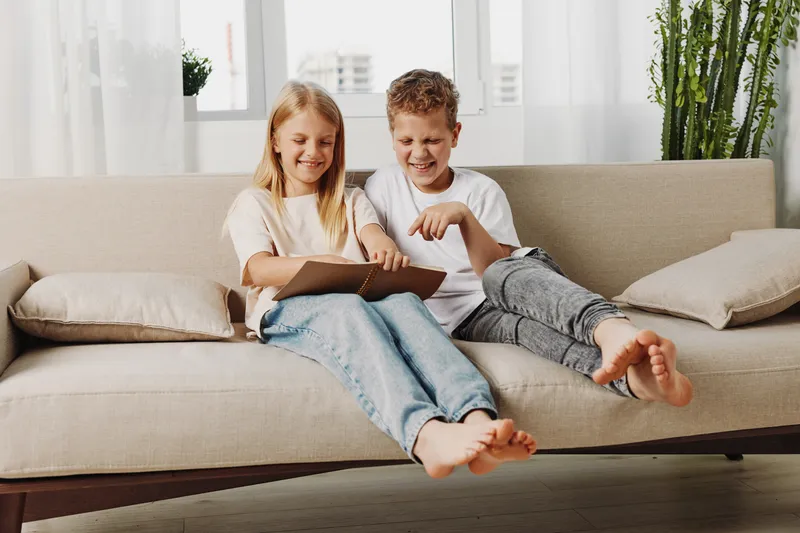 Two happy children sitting on a couch using a tablet together in a bright living room, enjoying digital entertainment and shared moments.