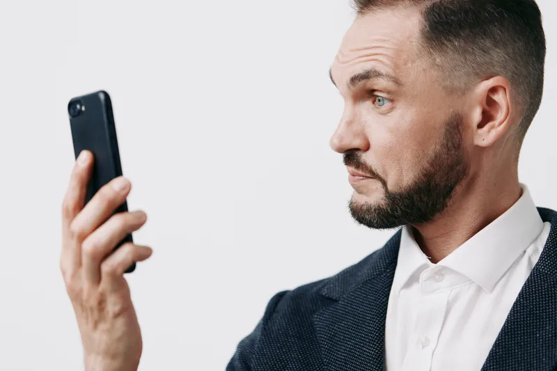 A focused business professional man examines a smartphone, captured against an isolated colored background with neutral tones, conveying concentration and intent.