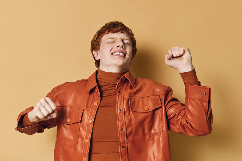 Young man with red hair wearing brown leather jacket smiling and celebrating, showing excitement and positive emotion on plain beige background.