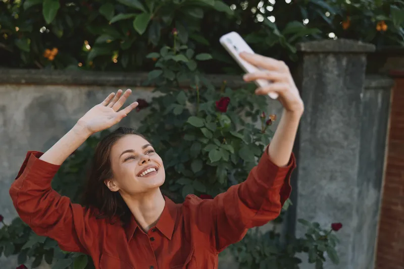 Happy woman takes a selfie outdoors, wearing a red shirt, smiling in a leafy garden, capturing a casual lifestyle moment against a natural backdrop in warm light