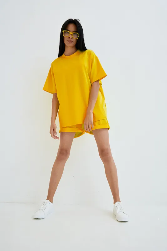 Woman wearing bright yellow casual outfit and sunglasses standing confidently against white background with relaxed pose and modern style.