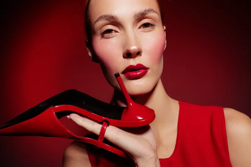 Close-up of a woman with red lipstick holding a red high heel shoe near her face. The red background and dress create a bold fashion and beauty concept with strong color contrast.