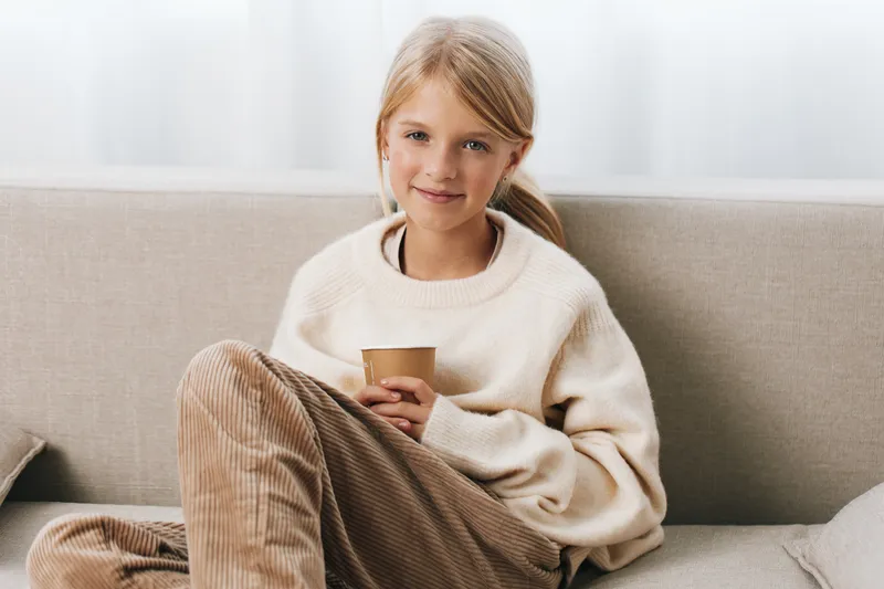 Young girl sits on a light sofa wearing a warm sweater, holding a cup with a calm smile, creating a cozy home scene with soft natural light and relaxed posture.