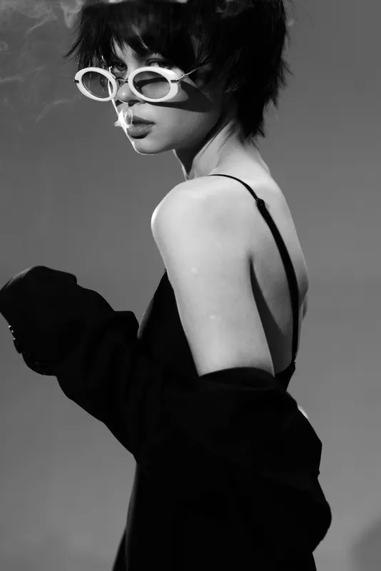 Black and white portrait of a stylish woman wearing sunglasses with a bare shoulder and layered hair, creating a moody and fashionable look.