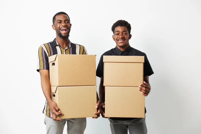 Two smiling black men holding cardboard boxes in a bright room, showcasing teamwork and collaboration in a business environment. Their joyful expressions evoke positivity.