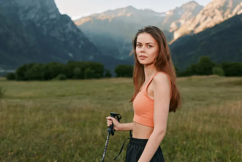 A confident woman stands in a wide alpine meadow with distant mountains. She wears an orange crop top and holds trekking poles, looking toward the camera with calm, focused energy.