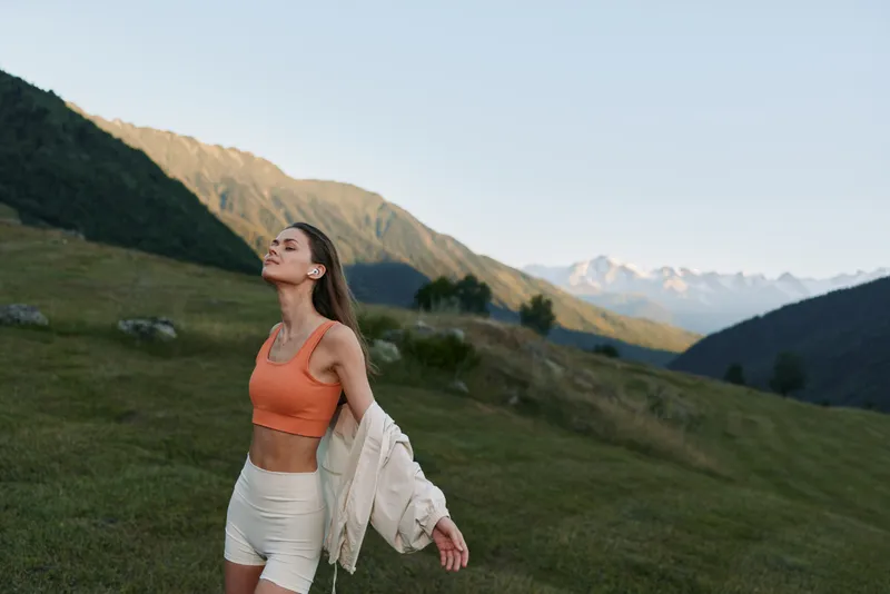 A young woman stands in a grassy hillside landscape, wearing an orange sports top and light shorts, with a jacket draped over her arm. She gazes toward the clear sky, enjoying fresh air and a
