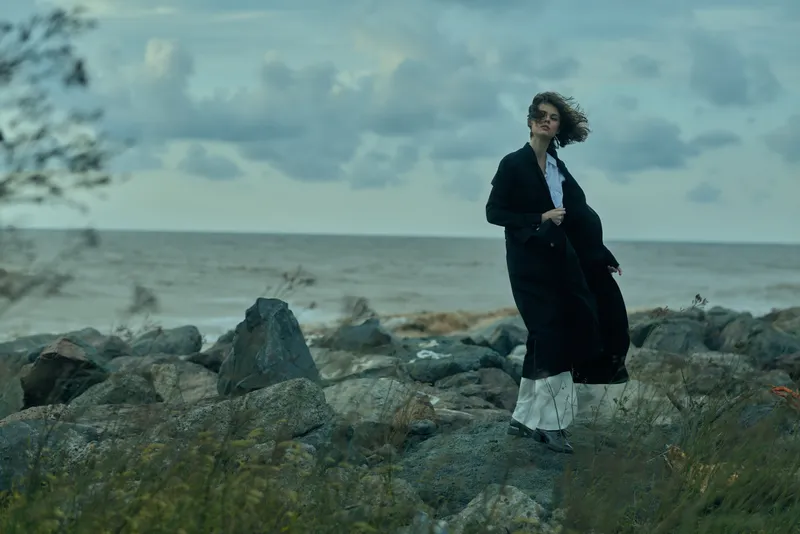 windswept coastline fashion, a dark coat on a figure by a rocky shoreline, moody ocean backdrop with dramatic styling and natural light for outdoor editorial use