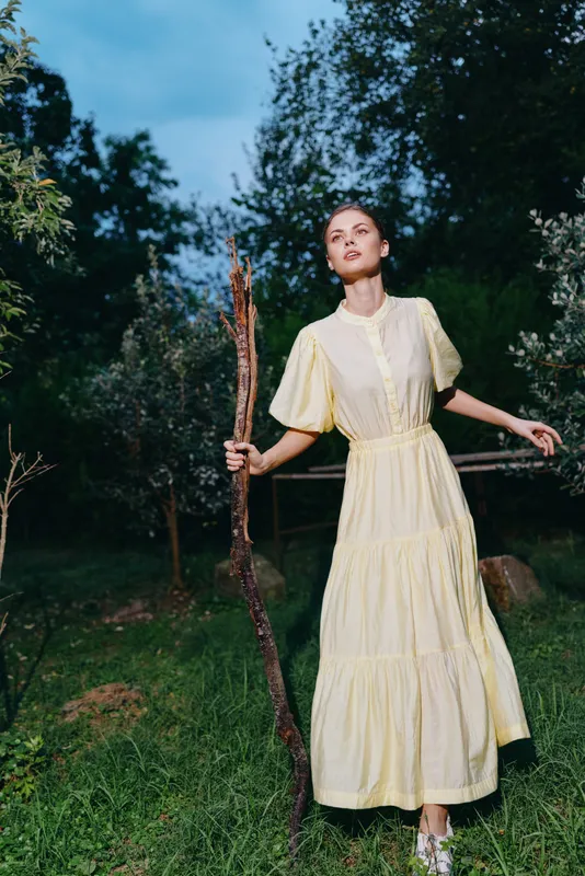 A woman in a pale yellow dress stands in a grassy countryside field holding a wooden staff, exuding serenity, elegance, and a graceful connection with nature