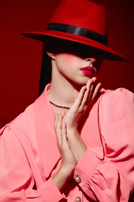 Elegant woman wearing a red hat and pink blazer poses with her hands near face, showcasing red lips and pearl necklace in creative fashion beauty shot on a red background.