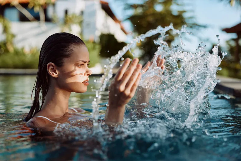 Young woman with dark brunette hair plays in a pool, splashing water toward her hands as sunlight reflects on the surface, conveying refreshment and carefree summer leisure.