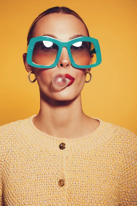 Young woman with large turquoise sunglasses blows a bubble gum bubble. She wears a yellow knitted sweater and gold hoop earrings against a bright yellow background. Fun and fashion concept.