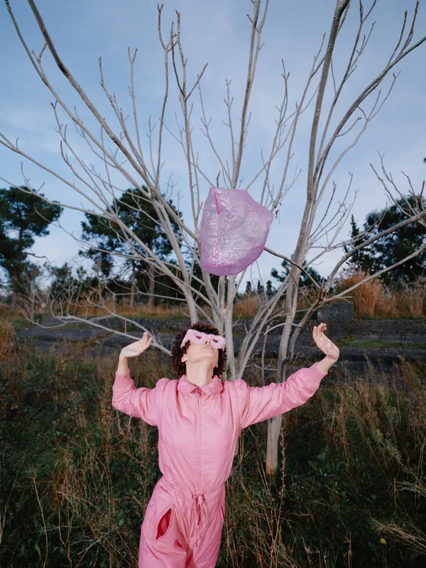 Pink jumpsuit worn by a person outdoors releases a pink heart into the air beside a leafless tree, a playful moment of joy and whimsy in an open field under clear sky.