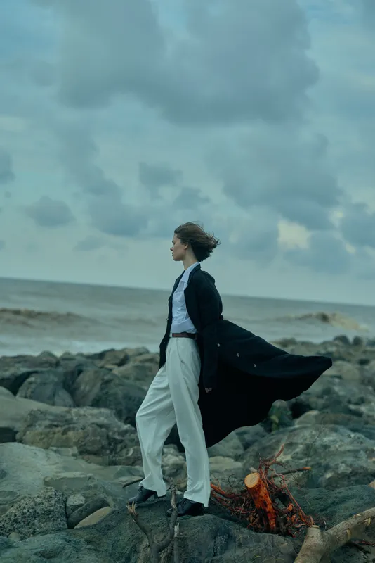 Fashionable woman in a tailored coat stands on rocky shore as wind billows the garment, dramatic coastline and open sea create a bold, atmospheric scene with dark clouds overhead and a sense of
