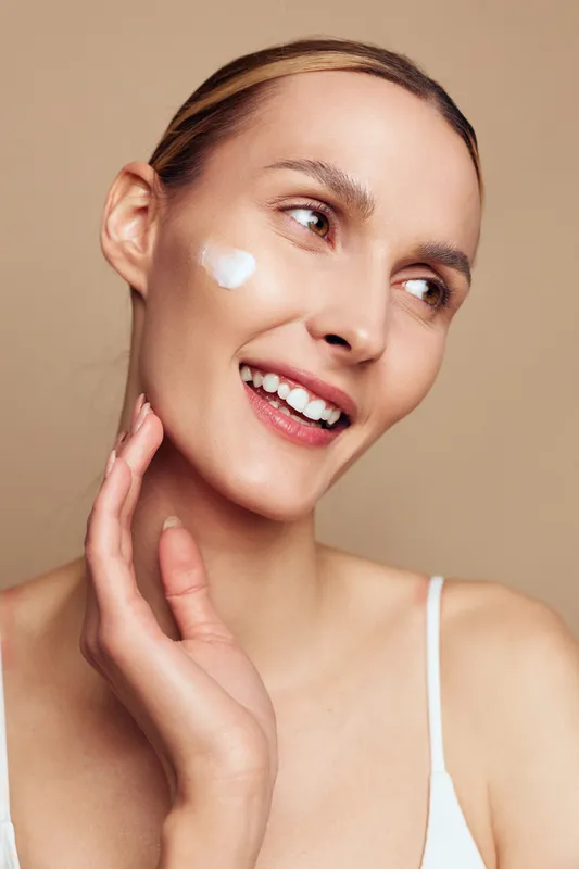 Young woman with clear skin applies white face cream on her cheek while smiling gently against a beige background in a skincare and self-care concept.